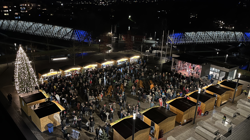 Weihnachtlich beleuchteter Marktplatz bei Dunkelheit. Holzhütten und eine Bühne sind aufgebaut. Viele Besucher sind auf dem Platz.