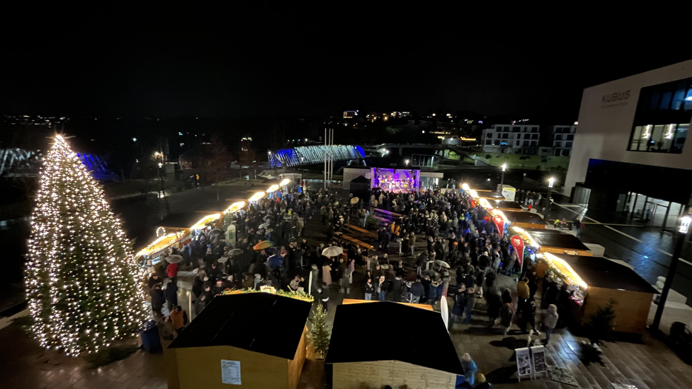 Weihnachtlich beleuchteter Marktplatz bei Dunkelheit. Holzhütten und eine Bühne sind aufgebaut. Viele Besucher sind auf dem Platz.