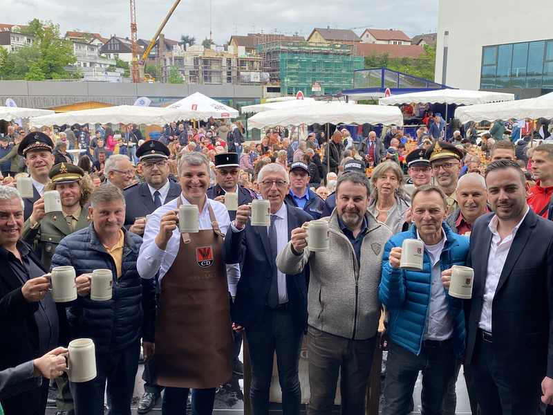 Mehrere Menschen mit Steinkrügen in der Hand, die sich vor einer Menschenmenge zuprosten.