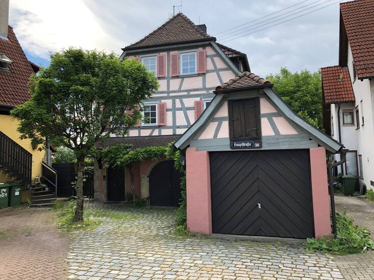 Ein altes Fachwerkhaus mit vorgebauter Garage. Vor dem haus steht ein mittelgroßer Baum.