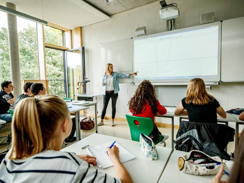 Klassenzimmer mit SChülern, an der Tafel steht die Lehrerin.