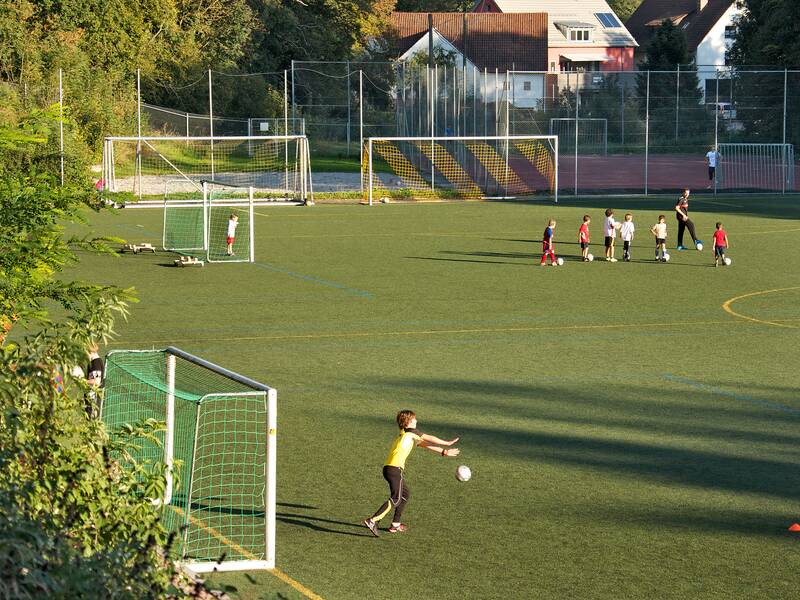 Kunstrasenplatz, Kinder spielen darauf Fußball.