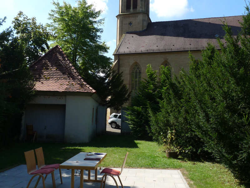 Zwei Stühle und ein Tisch im Freien, im Hintergrund sieht man eine Kirche.