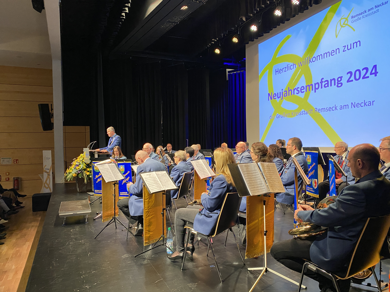 Musikverein sitzt auf einer Bühne, auf der linken Seite der Bühne steht ein Rednerpult, dahinter ein Redner. Auf der Leinwand im HIntergrund steht: Neujahrsempfang 2024.