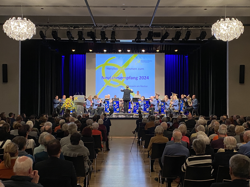 Stadthalle mit Menschen, die auf ihren Stühlen sitzen. Auf edr Bühne sitzt der Musikverein uns spielt ein Lied.