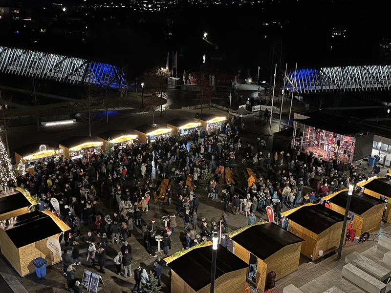 Weihnachtlich beleuchteter Marktplatz bei Dunkelheit. Holzhütten und eine Bühne sind aufgebaut. Viele Besucher sind auf dem Platz.